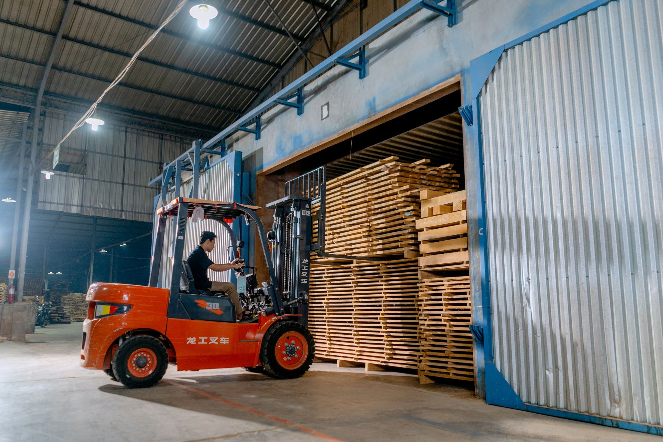 A forklift operator handling stacked lumber inside an industrial warehouse.