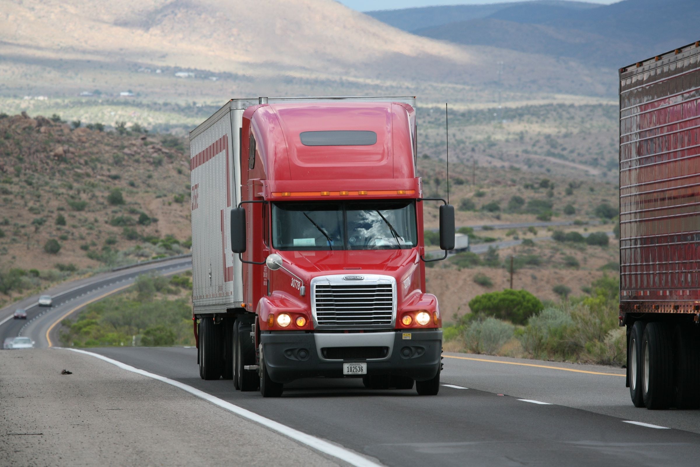 A red American semi-truck hauling a closed cargo trailer through a rugged mountain landscape.