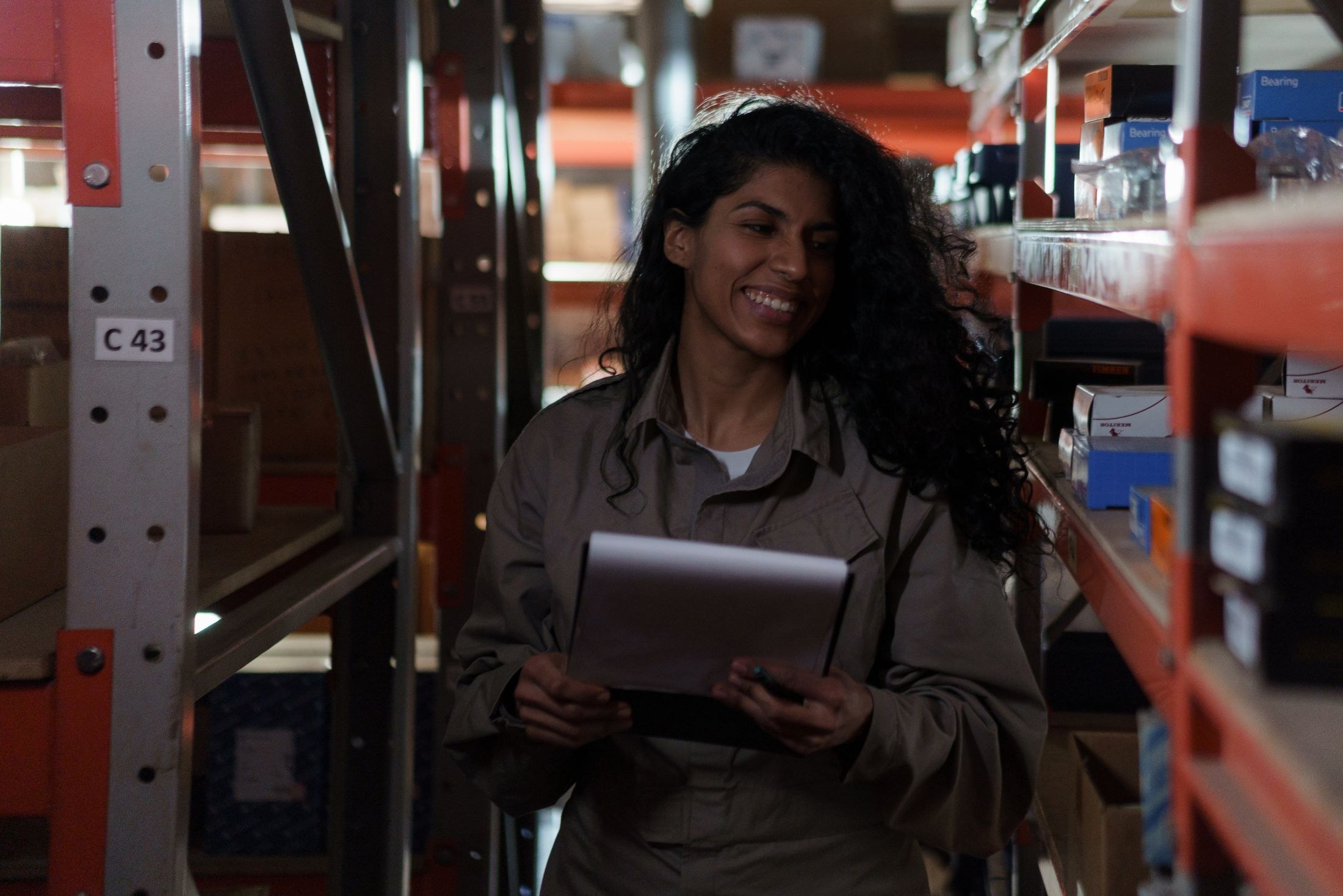 A woman holding a clipboard beside stocked shelving in a warehouse aisle.